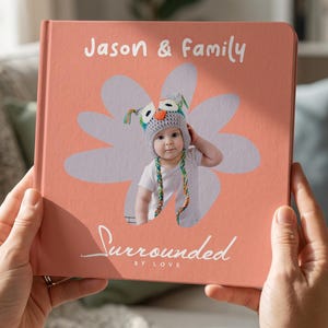 May include: A square photo book with a coral-colored cover featuring a white flower-shaped cutout. The cover displays the text "Jason & Family" and "Surrounded BY LOVE" with a central photograph of a baby wearing a gray owl-themed knit hat.