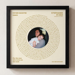 May include: A square black frame containing a cream-colored print with a circular arrangement of song lyrics surrounding a central photograph of a person holding a child. The text includes FATHER DAUGHTER DANCE, STEPHANIE & DAD, AUGUST 24, 2024, For the man who loves me the most, and ZAC BROWN BAND.