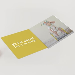 May include: Open photo book with a yellow cover and a photo of a baby wearing an owl hat. The cover reads "Hi! I'm Jason. This is my family!" in white text. The photo shows the baby in a wire basket.