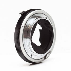 May include: A silver and black camera lens adapter with a K/R marking. The adapter has a smooth, silver outer ring and a black inner ring with a circular opening.