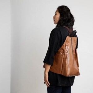 Puede incluir: Una mujer con una camisa negra y una mochila de cuero marr&oacute;n. La mochila tiene una cremallera y una solapa.