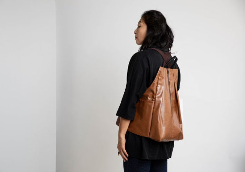 Puede incluir: Una mujer con una camisa negra y una mochila de cuero marr&oacute;n. La mochila tiene una cremallera y una solapa.