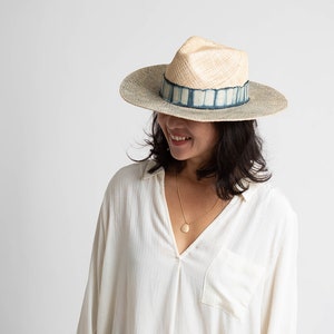 May include: A woman wearing a white shirt and a wide-brimmed straw hat with a blue and white patterned band. The hat has a slightly pointed crown.