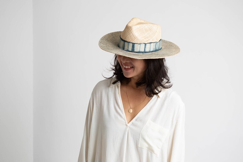 May include: A woman wearing a white shirt and a wide-brimmed straw hat with a blue and white patterned band. The hat has a slightly pointed crown.