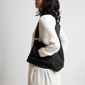 May include: A woman wearing a white shirt and brown trousers is carrying a black leather rucksack.