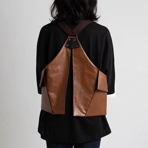 Puede incluir: Una mochila de cuero marr&oacute;n con un dise&ntilde;o &uacute;nico. La bolsa tiene forma triangular con dos bolsillos delanteros y correas marrones. La persona de la imagen lleva una camisa negra y vaqueros azules.