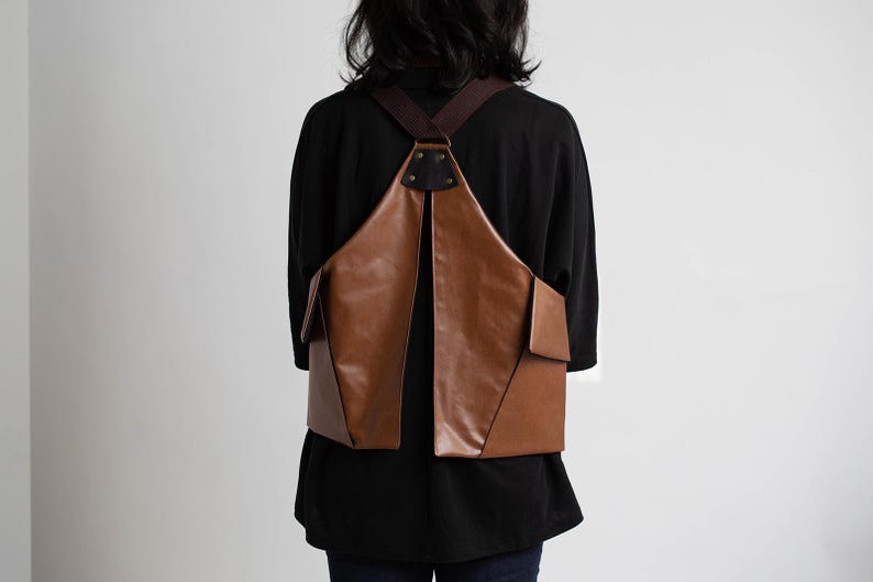 Puede incluir: Una mochila de cuero marr&oacute;n con un dise&ntilde;o &uacute;nico. La bolsa tiene forma triangular con dos bolsillos delanteros y correas marrones. La persona de la imagen lleva una camisa negra y vaqueros azules.