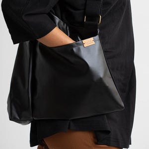 Puede incluir: Bolso mensajero negro con correa para el hombro. El bolso est&aacute; hecho de un material brillante y liso y tiene forma rectangular. Una peque&ntilde;a etiqueta rectangular est&aacute; adherida al bolso. El bolso lo lleva una persona que viste una camisa negra.