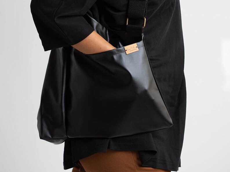 Puede incluir: Bolso mensajero negro con correa para el hombro. El bolso est&aacute; hecho de un material brillante y liso y tiene forma rectangular. Una peque&ntilde;a etiqueta rectangular est&aacute; adherida al bolso. El bolso lo lleva una persona que viste una camisa negra.