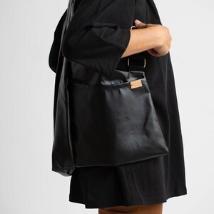 May include: A black leather tote bag with a brown leather label. The bag is being worn by a person.