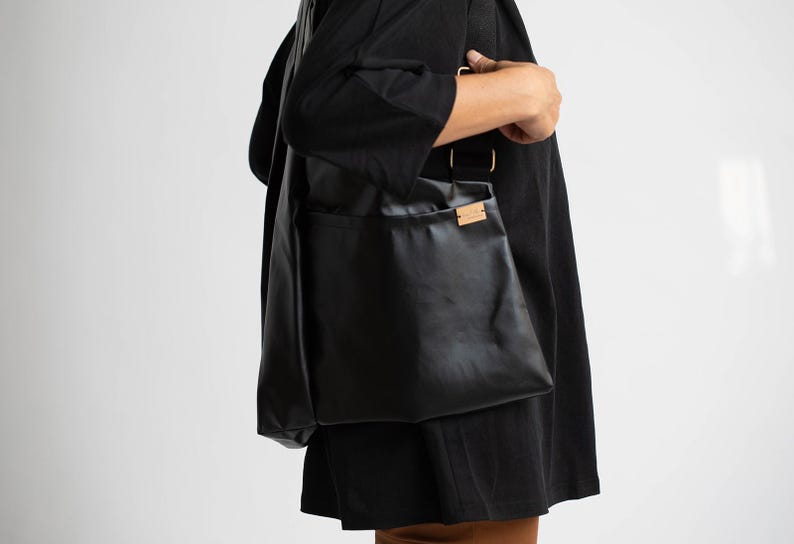 May include: A black leather tote bag with a brown leather label. The bag is being worn by a person.