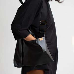 May include: A black leather tote bag with a long adjustable strap. The bag has a small pocket on the front with a brown leather label. The bag is being worn by a person wearing a black shirt and brown trousers.