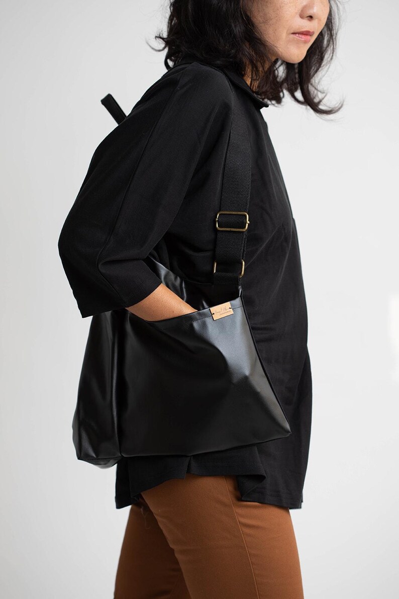 May include: A black leather tote bag with a long adjustable strap. The bag has a small pocket on the front with a brown leather label. The bag is being worn by a person wearing a black shirt and brown trousers.