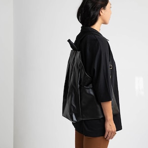 May include: A black leather backpack with a flap closure and adjustable straps. The backpack is being worn by a person wearing a black shirt and brown trousers.