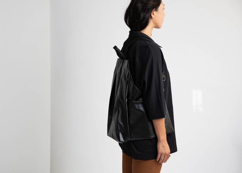 May include: A black leather backpack with a flap closure and adjustable straps. The backpack is being worn by a person wearing a black shirt and brown trousers.