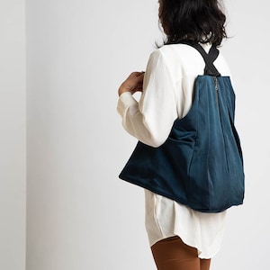 Puede incluir: Una mochila azul turquesa con correas negras y cierre de cremallera. La mochila la lleva puesta una persona que lleva una camisa blanca y pantalones marrones.