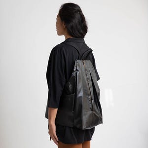 May include: A black, pleated, zippered backpack with a gold zipper pull. The backpack is being worn by a person wearing a black long-sleeved shirt.