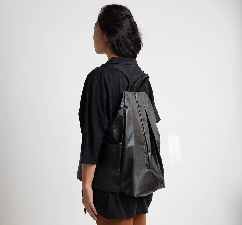 May include: A black, pleated, zippered backpack with a gold zipper pull. The backpack is being worn by a person wearing a black long-sleeved shirt.