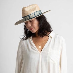 May include: A woman wearing a white shirt with a v-neck and a straw hat with a blue and white patterned band. She is wearing a gold necklace with a pendant.