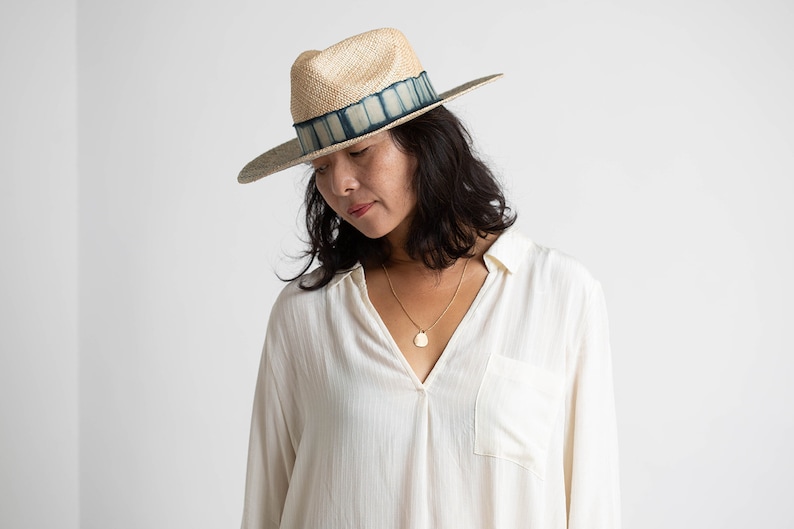 May include: A woman wearing a white shirt with a v-neck and a straw hat with a blue and white patterned band. She is wearing a gold necklace with a pendant.