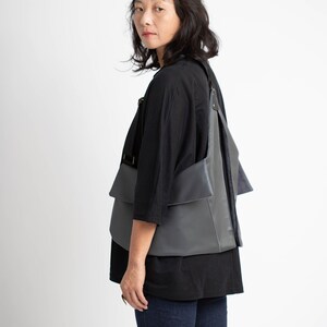 Puede incluir: Bolso de hombro de cuero gris con un dise&ntilde;o &uacute;nico en capas. El bolso tiene forma rectangular con m&uacute;ltiples bolsillos y correas ajustables. Se lleva sobre una parte superior negra y sobre un fondo blanco.