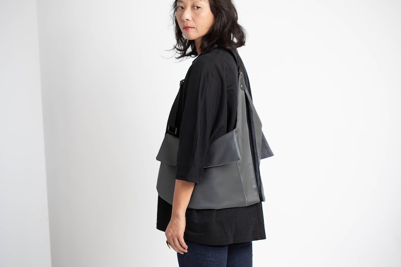 Puede incluir: Bolso de hombro de cuero gris con un dise&ntilde;o &uacute;nico en capas. El bolso tiene forma rectangular con m&uacute;ltiples bolsillos y correas ajustables. Se lleva sobre una parte superior negra y sobre un fondo blanco.