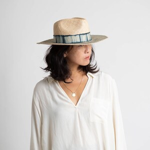 May include: A woman wearing a white shirt and a wide-brimmed straw hat with a blue and white patterned band. The hat has a slightly rounded crown and a wide brim.
