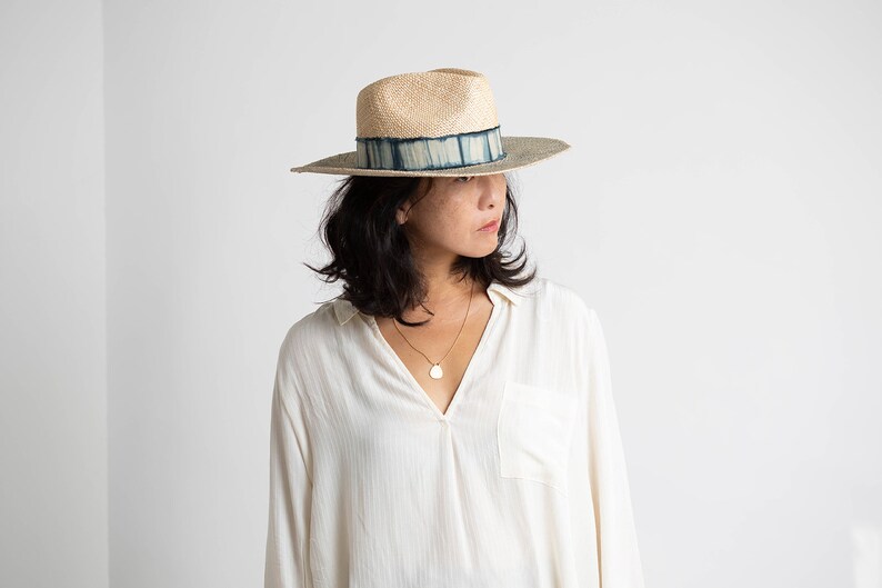 May include: A woman wearing a white shirt and a wide-brimmed straw hat with a blue and white patterned band. The hat has a slightly rounded crown and a wide brim.