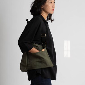 Pu&ograve; includere: Una donna indossa una camicia nera e jeans blu scuro e porta una borsa a tracolla in tela verde con una tracolla nera e una tasca in pelle marrone.