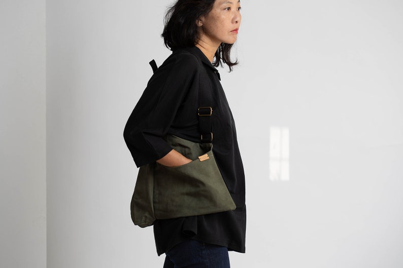 Pu&ograve; includere: Una donna indossa una camicia nera e jeans blu scuro e porta una borsa a tracolla in tela verde con una tracolla nera e una tasca in pelle marrone.