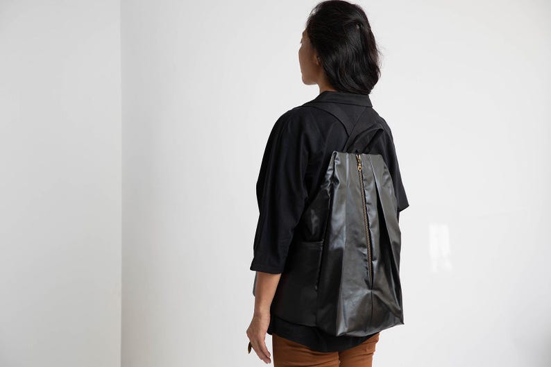 May include: A person wearing a black shirt and brown trousers with a black rucksack. The rucksack has a zipper and a pleated design.