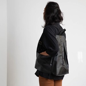 May include: A person wearing a black shirt and brown trousers with a black leather rucksack. The rucksack has a zip and a flap.