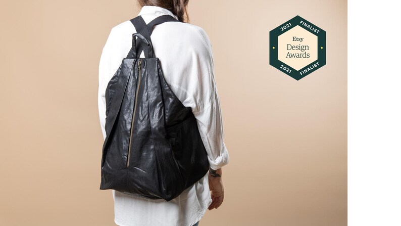Puede incluir: Una mochila de cuero negro con cierre de cremallera y correas ajustables. La mochila la lleva puesta una persona y es finalista de los Etsy Design Awards 2021.