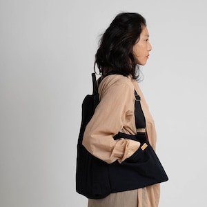 Puede incluir: Una persona lleva una camisa de color beige con una mochila negra. La mochila tiene una correa que se coloca sobre el hombro y un bolsillo en la parte delantera.