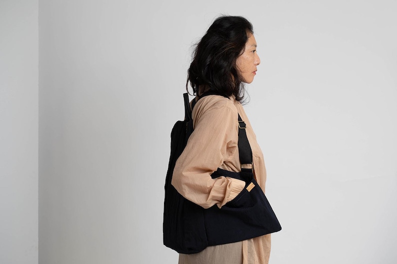 Puede incluir: Una persona lleva una camisa de color beige con una mochila negra. La mochila tiene una correa que se coloca sobre el hombro y un bolsillo en la parte delantera.