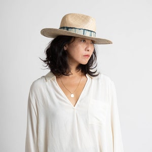 May include: A woman wearing a white button-down shirt with a light brown wide-brimmed hat with a blue and white patterned band. She is wearing a gold necklace with a round pendant.
