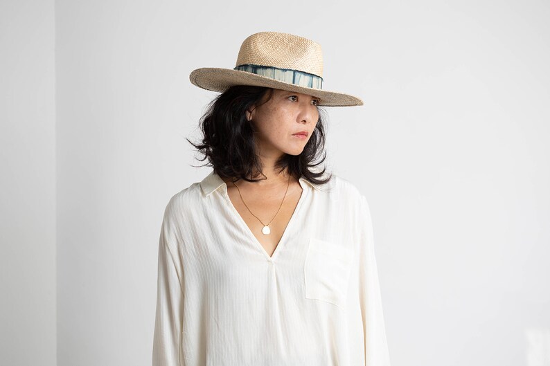 May include: A woman wearing a white button-down shirt with a light brown wide-brimmed hat with a blue and white patterned band. She is wearing a gold necklace with a round pendant.