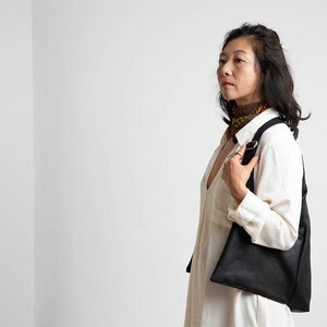 May include: A woman wearing a white shirt and a patterned scarf is holding a black leather tote bag.