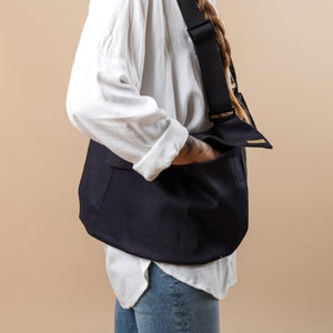 Puede incluir: Un bolso bandolera de lona negra con una correa larga ajustable. El bolso tiene cierre de solapa y un pequeño bolsillo en la parte delantera.