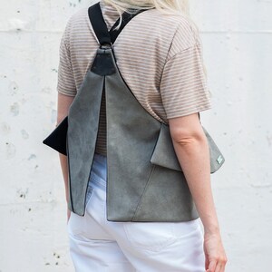 Puede incluir: Una mochila de ante gris con correas y detalles en negro. La bolsa tiene un dise&ntilde;o geom&eacute;trico &uacute;nico con cierre de solapa y un peque&ntilde;o bolsillo. La persona lleva una camisa a rayas y pantalones blancos.