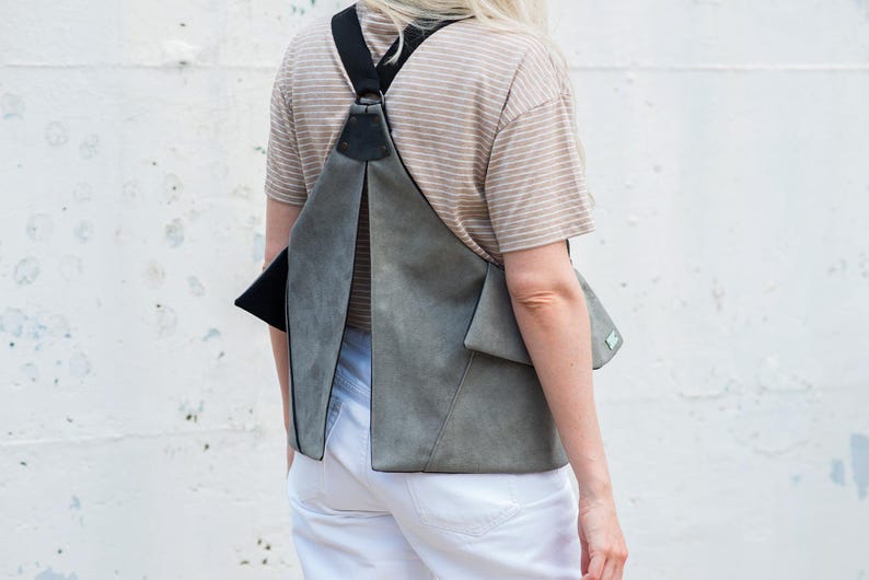 Puede incluir: Una mochila de ante gris con correas y detalles en negro. La bolsa tiene un dise&ntilde;o geom&eacute;trico &uacute;nico con cierre de solapa y un peque&ntilde;o bolsillo. La persona lleva una camisa a rayas y pantalones blancos.