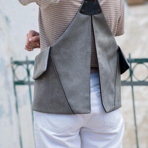 Puede incluir: Un bolso de hombro gris con un dise&ntilde;o geom&eacute;trico &uacute;nico. El bolso tiene un bolsillo con solapa y una correa de cuero negro. La persona lleva una camisa a rayas y pantalones blancos. El bolso est&aacute; hecho de un material suave, similar al ante.