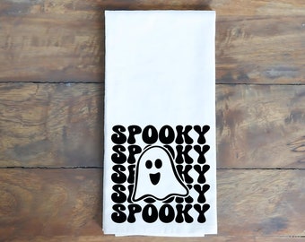 May include: A white rectangular kitchen towel featuring a repeating black graphic pattern of the word SPOOKY in a wavy font, with a central illustration of a smiling ghost. The design is centered on the lower portion of the fabric.