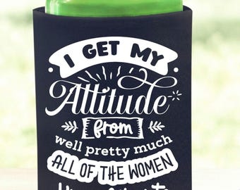 May include: Black drinks cooler with white text: "I GET MY Attitude from well pretty much ALL OF THE WOMEN I'm related to". A green can sits inside. The cooler rests on a wooden coaster.