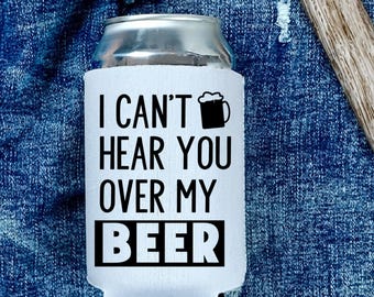 May include: A white foam beverage insulator sleeve for a metal can. The sleeve features black text that reads I CAN'T HEAR YOU OVER MY BEER, accompanied by a small black graphic of a beer mug with foam at the top, centred on the front surface.