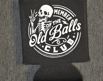 May include: A black foam beverage insulator featuring a white graphic of a skeleton making a peace sign. The text reads MEMBER OF THE Old Balls CLUB in a circular design. The graphic is printed in white ink on the front of the black fabric sleeve.