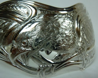 May include: A silver bracelet with a detailed design of two eagles with their wings spread out. The eagles are facing each other and their heads are close together. The bracelet is made of a smooth, polished metal.