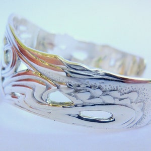 May include: Silver bracelet with an intricate, ornate design. The bracelet is open and has a smooth, polished finish.