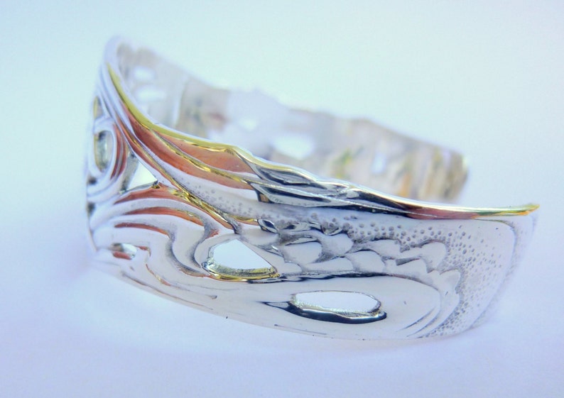 May include: Silver bracelet with an intricate, ornate design. The bracelet is open and has a smooth, polished finish.