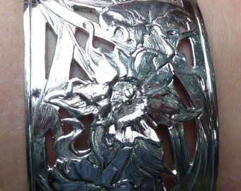 May include: A silver-colored cuff bracelet with a floral design. The bracelet features a wide band with cut-out patterns and detailed flower and leaf motifs. The bracelet is approximately 2.5 inches wide.